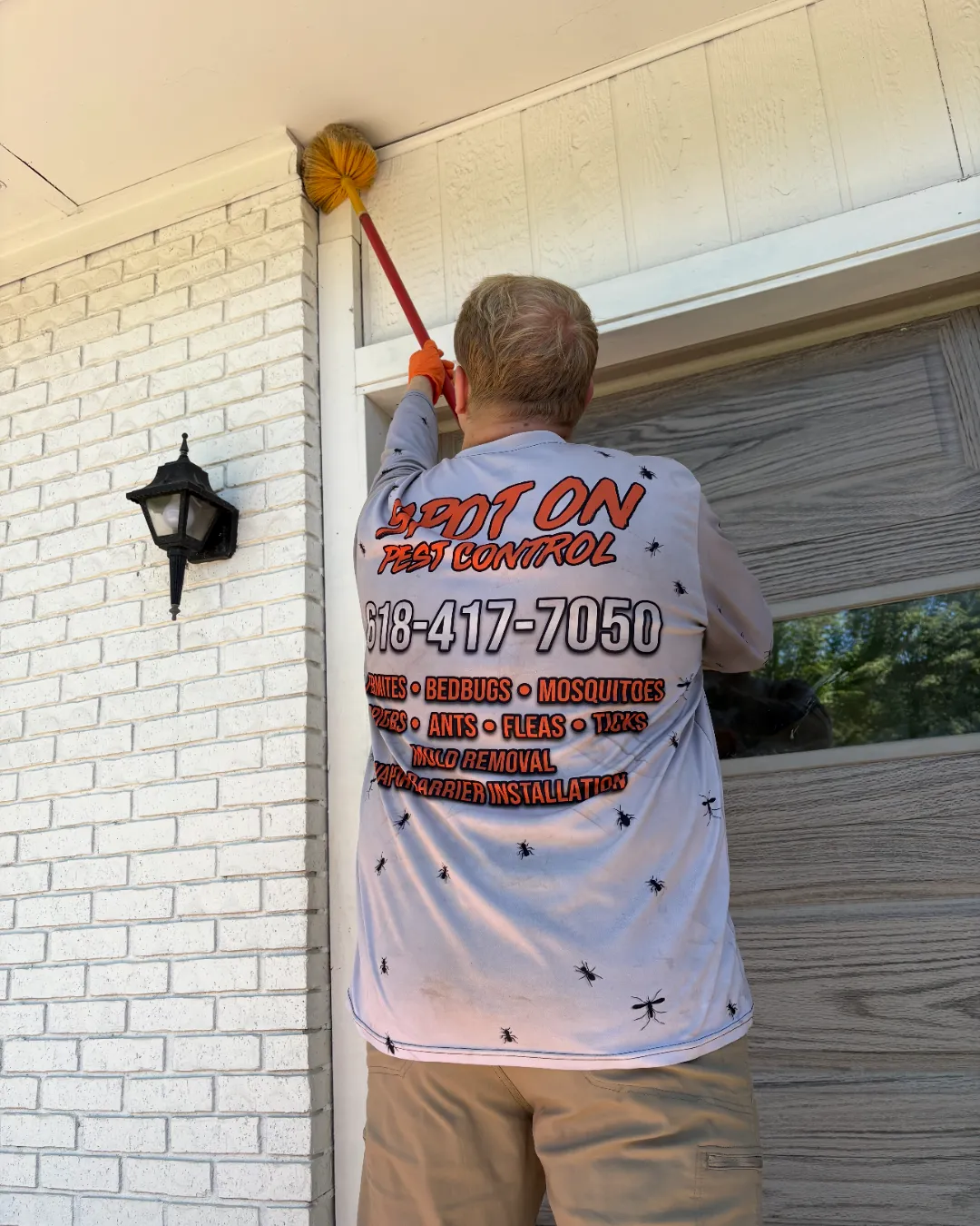 A Spot On technician, in a shirt printed with the company phone number, treating the eaves of a brick home.